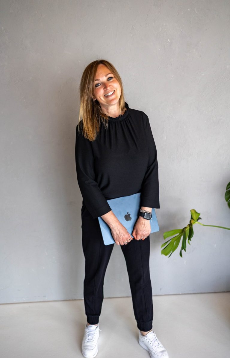 Sabine Welsch mit blonden Haaren in schwarzer Kleidung hält einen Laptop und lächelt.